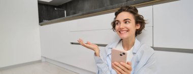 Mutlu genç bir kadının portresi, yepyeni mutfağını gösteriyor, yere oturuyor, mobilyaları gösteriyor ve gülümsüyor, akıllı telefondaki video sohbetleri. Teknoloji ve yaşam tarzı kavramı