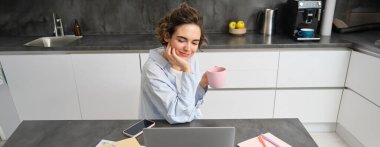 Güzel esmer bir kız, evden online kurs okuyan bir öğrenci, uzaktan kumanda üzerinde çalışan, dizüstü bilgisayara bakan, bir fincan kahveyle mutfakta oturan.