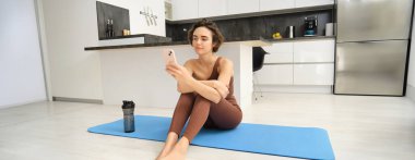 Spor giyim sektöründe fitness kızı portresi, cep telefonuna bakmak, evden antrenman yapmak, akıllı telefon uygulamasında egzersiz talimatları aramak..