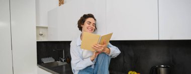 Yaşam tarzı ve modern insan konsepti. Genç bir kadın günlük okuyor, notlarına bakıyor ve gülüyor, evde eğleniyor, mutfak tezgahında oturuyor ve gülüyor..