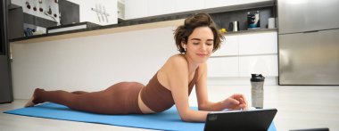 Genç fitness eğitmeni, kadın yoga yapıyor, tablete bakıyor, çevrimiçi video pilates dersi izliyor, meditasyon eğitimi alıyor, kablosuz kulaklıkla dinliyor, lastik paspasın üzerinde yatıyor, evde spor salonunda..