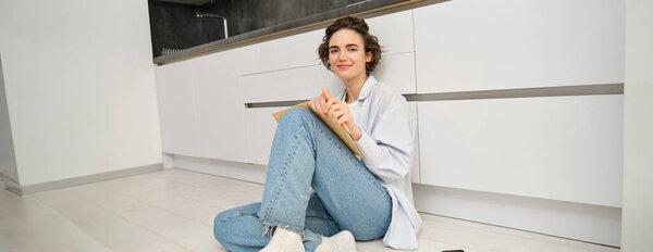 Portrait of woman writing down her thoughts in diary, planner or journal, holding notebook on her laps, sitting on floor in kitchen and studying.