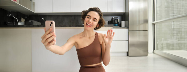 Изображение молодой фитнес-девушки блоггер, записывает ее тренировки дома, чаты в Интернете во время занятий йогой в светлой комнате. Женщина со смартфоном, присоединяющаяся к дистанционному обучению йоге через мобильный телефон.