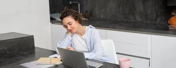Image of young productive woman works from home, does distance learning, learns online course on her laptop in kitchen, writes down information, watches webinar, attends work meeting remotely.