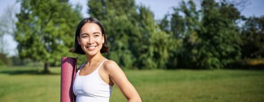 Lastik paspaslı gülümseyen fitness kızı, spor ve spor için üniforma giyerek parkta duruyor, çimenlikte yoga yapıyor..