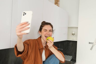 Güzel bir genç kadın, yaşam tarzı blogcusu kendini videoya kaydediyor, elma yerken selfie çekiyor, mutfak tezgahında oturuyor, akıllı telefon kullanıyor..