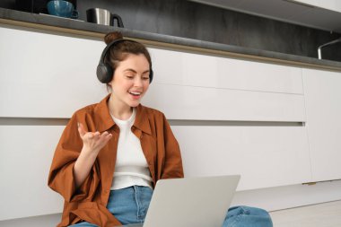 Serbest çalışan genç bir kadın online sohbete katılıyor, evde okuyan bir öğrenci, yerde dizüstü bilgisayar ve kulaklıkla oturuyor..