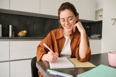 Modern genç bir kadının gözlüklü portresi, evde oturan ve ödev yapan öğrenci, not defterine bilgi yazan ve gülümseyen, proje üzerinde çalışan..