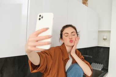 Genç karizmatik kız mutfakta fotoğraf için selfie çekiyor, cep telefonunu uzatarak tutuyor..