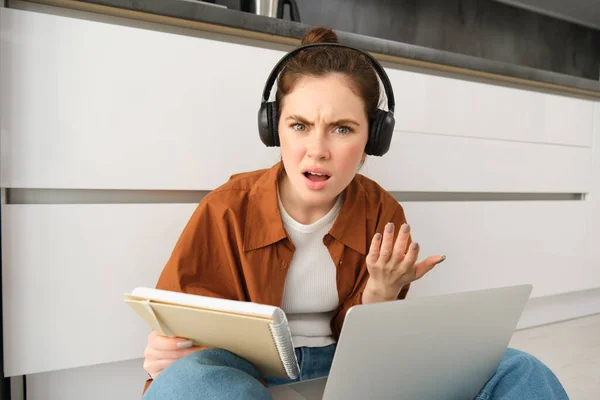 Portrait of annoyed and confused woman in headphones, sits on floor with laptop and notebook, looks frustrated, has issues in doing homework or studying.