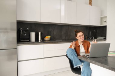 Evden çalışan, dizüstü bilgisayarla mutfakta oturan, çevrimiçi toplantıya katılan ve uzaktan kumandada müşteriyle konuşan, bilgisayar başında sohbet eden şık bir kadının portresi..