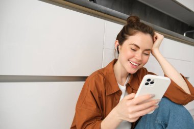Akıllı telefonlu, mesajlaşan, cep telefonu kullanan, mutfak zemininde oturan, kablosuz kulaklıkla müzik dinleyen tasasız, güler yüzlü bir kadın..