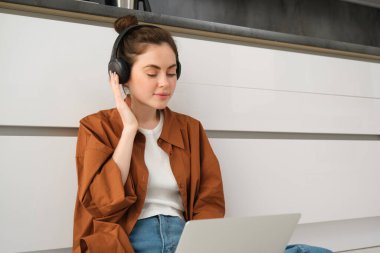 Genç bir kadın, laptopta ödev yapıyor, yerde oturuyor, kulaklıkla müzik dinliyor ve evden çalışıyor..
