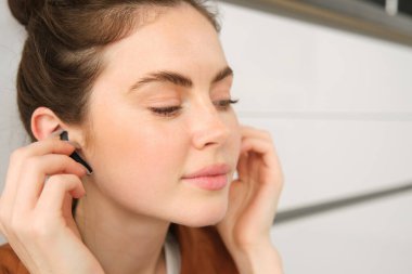 Modern esmer kız imajı, mutfak zemininde oturuyor, kablosuz kulaklıkla müzik dinliyor, kulaklık tutuyor ve elinde çanta, yan yana bakıyor..