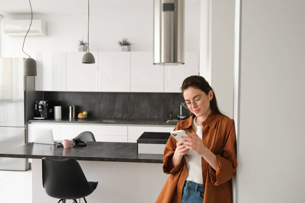 Portrait of young woman at home, checking news or messages on smartphone, online shopping, confirm delivery on mobile phone app, leaning on wall, standing in living room.