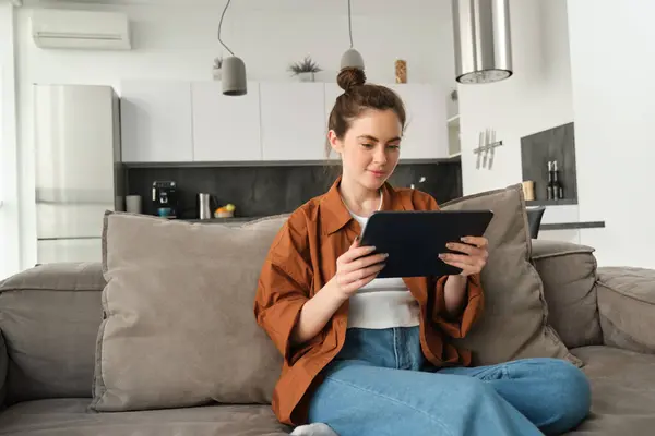 stock image Portrait of young woman sitting on couch in living room, watching movies on digital tablet, reading on device, enjoying weekend at home.