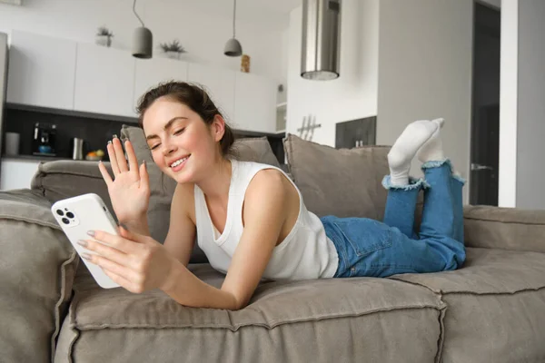 Happy cute female model, lying on couch, waving hand at smartphone, saying hi, connects to video call, resting at home and talking to friend.