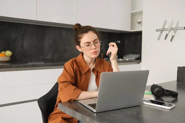 Portrait of woman with serious face, sitting in front of computer, using laptop, working from home, looking at screen with thoughtful face.