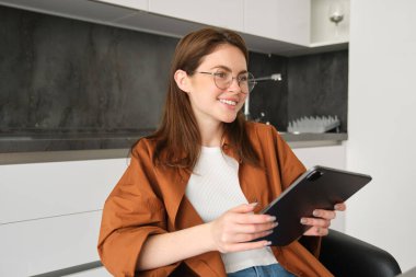 Genç bir öğrencinin portresi, evde okuyan bir kadın, uzaktan çalışıyor, mutfağında iş yeri kuruyor, dijital tabletle sandalyeye oturuyor, gözlük takıyor..