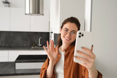 Gülümseyen genç bir kadının portresi cep telefonuyla evde duruyor, akıllı telefona el sallıyor, online sohbete bağlanıyor, biriyle konuşuyor..