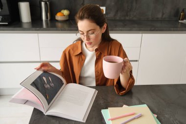 Hafta sonunun tadını çıkaran, kitap okuyan, evde çay içen, gözlük takan ve günlük giysiler giyen genç bir kadının portresi..