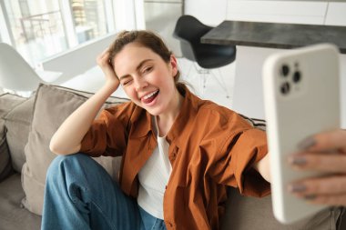 Güzel, mutlu genç kadın modelin portresi, akıllı telefonlu koltukta oturuyor, cep telefonuyla selfie çekiyor, gülen surat yapıyor..