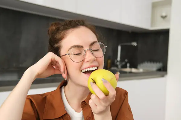 Portrait of smiling, modern woman in glasses, sitting at home and eating green apple, having a lunch break, biting fruit.