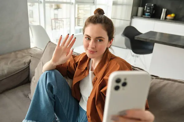 Happy woman is sitting at home with smartphone, online chatting, waving hand at mobile phone, saying hello, talking to friends using application, sitting on sofa at home.