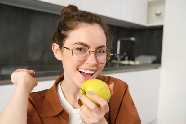 Close-up portrait of brunette woman at home, wearing glasses, eating apple in the kitchen and smiling, biting fruit.