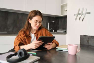 Gözlüklü güzel bir genç kadının portresi, dijital tablet okuyor, video izliyor, uzaktan çalışıyor, kulaklıklar, kağıtlar ve defterlerle mutfakta oturuyor..