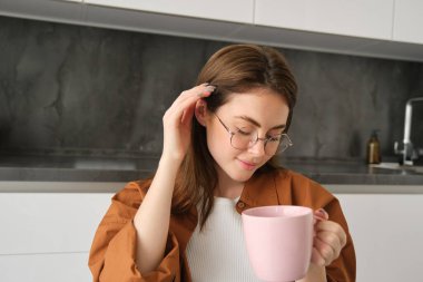 Gözlüklü, tatlı ve hassas genç bir kadının portresi, mutfakta oturuyor, çay içiyor, fincanı tutuyor, saçını kulağının arkasına sıkıştırıyor ve gülümsüyor..
