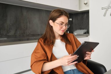 Gözlüklü esmer bir kadının portresi, mutfağında dijital tabletle oturuyor, aletleri okuyor, internetten video izliyor, uzaktan kumandayla çalışıyor..