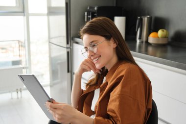 Güzel bir genç kadının portresi, evde dijital tablet kullanan bir öğrenci, mutfakta oturuyor, gözlük takıyor ve gülümsüyor, evden çalışıyor, internetten çalışıyor..