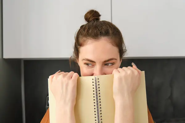 Cute woman hides face behind notebook, looking aside, smiling with eyes.