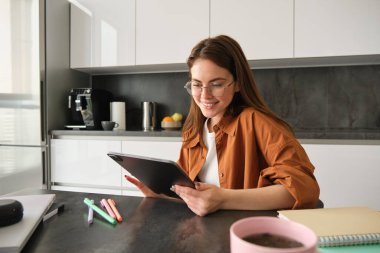 Genç bir öğrencinin portresi, evde okuyan bir kadın, uzaktan çalışıyor, mutfağında iş yeri kuruyor, dijital tabletle sandalyeye oturuyor, gözlük takıyor..