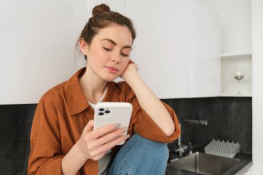 Modern stil kızı görüntüsü mutfak tezgahında telefonuyla oturuyor, cep telefonuna bakıyor, mesajları kontrol ediyor, akıllı telefondaki sosyal medya uygulamasını kaydırıyor..