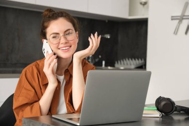 Evden çalışan güzel gülümseyen bir kadının portresi, cep telefonuyla konuşan, müşteriyi arayan, serbest çalışan bir iş kadını, dizüstü bilgisayar ve akıllı telefon kullanarak mutfakta iş yeri kuruyor..