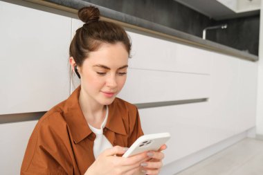 Gülümseyen güzel bir kadının portresi mutfak zemininde oturuyor, cep telefonuyla sosyal medyayı kaydırıyor, kablosuz kulaklıkla müzik dinliyor..