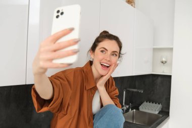 Yeni akıllı telefonunda selfie çeken genç mutlu bir kadın gülümseyen, sevimli bir yüz ifadesi olan ve mutfakta oturan fotoğraflara poz veriyor..
