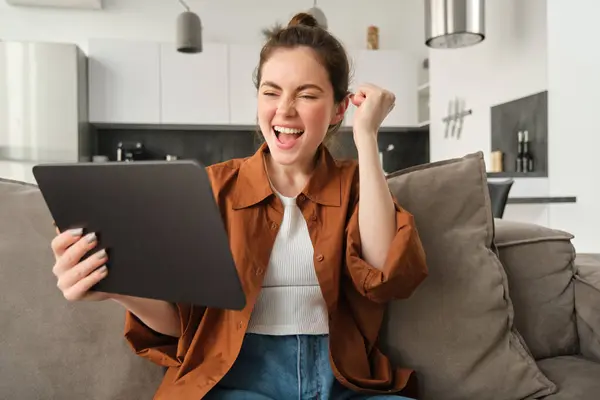 Happy, enthusiastic young woman winning on video game, looking at digital tablet and celebrating victory, triumphing and laughing, sitting on couch at home.