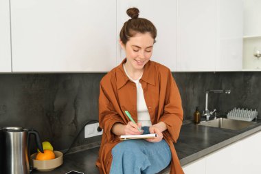 Mutfak tezgahında genç bir kadının portresi, defterle oturuyor, yaratıcı fikirlerini kağıda yazıyor, notlar alıyor, alışveriş listesi hazırlıyor..