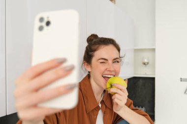 Mutlu, gülümseyen genç kadın portresi kendini kaydediyor, mutfakta elma yerken selfie çekiyor, akıllı telefon uygulaması kullanıyor, cep telefonuyla fotoğraf çekiyor..