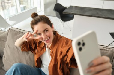 Pozitif ve mutlu genç bir kadın akıllı telefondan selfie çekiyor, kanepede oturuyor, huzur gösteriyor, v-işareti işareti işareti yapıyor..
