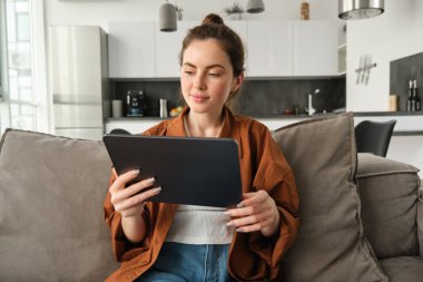 Oturma odasında kanepede oturan genç bir kadının portresi dijital tablette film izliyor, cihazda okuyor, hafta sonunun keyfini çıkarıyor..