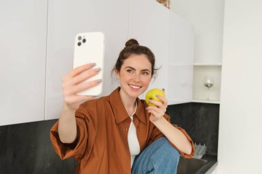 Mutlu, gülümseyen genç kadın portresi kendini kaydediyor, mutfakta elma yerken selfie çekiyor, akıllı telefon uygulaması kullanıyor, cep telefonuyla fotoğraf çekiyor..