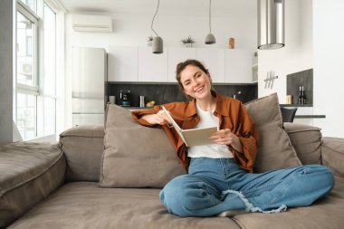 Genç bir kadın kanepede oturuyor, notlarını okuyor, not defteri tutuyor, sınava çalışıyor, oturma odasında öğrenci düzeltmeleri yapıyor..