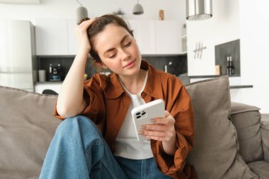 Güzel bir genç kadının portresi cep telefonuyla haber okuyor, akıllı telefon uygulaması kullanıyor, ekrana bakıyor ve gülümsüyor, evinde kanepede oturuyor..