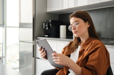 Ev ve eğlence konsepti. Güzel genç bir kadının portresi dijital tablette okuyor, mutfakta oturuyor, uzaktan kumandayla çalışıyor..