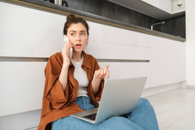 Endişeli, endişeli yüzlü bir kadın, bir telefon görüşmesi yapıyor, dizüstü bilgisayarla yerde oturuyor ve telefonda zor bir konuşma yapıyor..