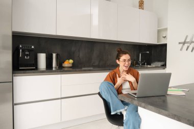 Evden çalışmak, uzaktan iş kavramı. Genç bir kadın mutfakta oturuyor, dizüstü bilgisayar kullanıyor, görüntülü sohbete katılıyor, çevrimiçi dersler veriyor, bilgisayarda müşteriyle konuşuyor..